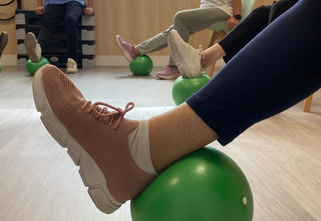 Personas mayores sentadas trabajando el equilibrio con un pie apoyado sobre un balón, en la clase de ejercicio terapéutico de Punto Fisio en Ciudad Lineal